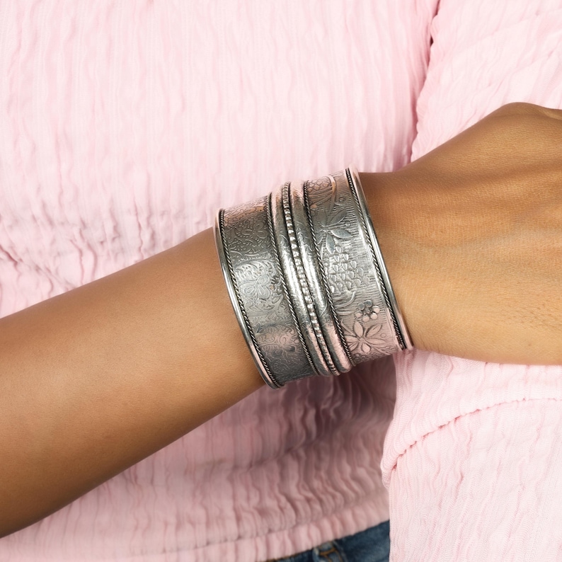 May include: Silver-toned cuff bracelets with intricate floral and geometric patterns. The bracelets are wide and stacked, with detailed engravings and a textured finish. The jewelry is worn on the wrist, showcasing the craftsmanship and design.