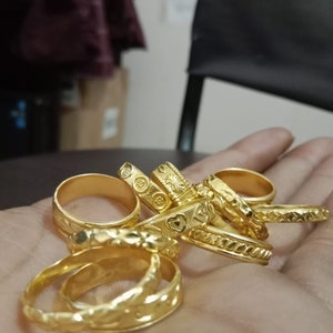 May include: A collection of gold rings with various designs, including floral, heart, and geometric patterns. The rings are displayed in a hand, showcasing their polished surfaces and intricate details. The rings vary in size and style.