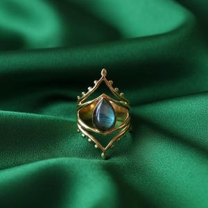 May include: A gold-colored ring with a teardrop-shaped, blue-gray gemstone. The ring has an ornate, openwork design with a crown-like top and decorative elements. The ring is set against a dark green fabric background.