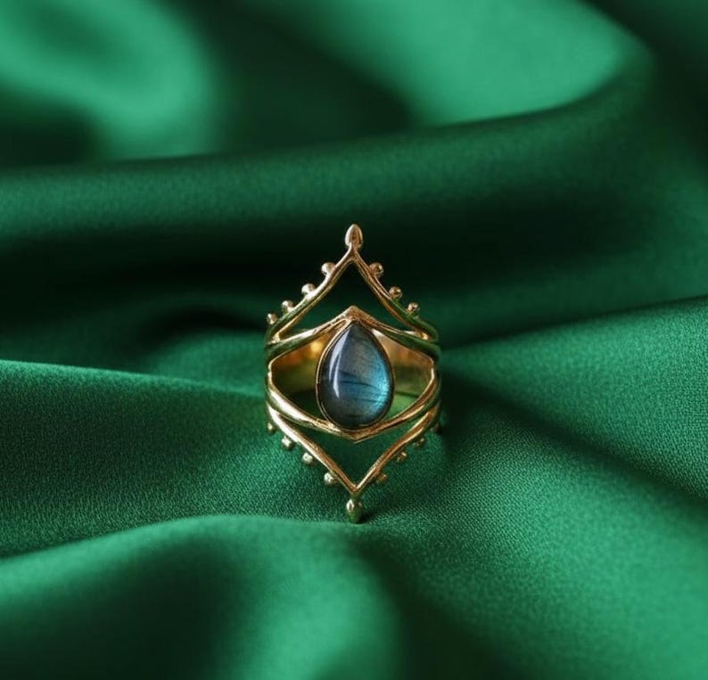 May include: A gold-colored ring with a teardrop-shaped, blue-gray gemstone. The ring has an ornate, openwork design with a crown-like top and decorative elements. The ring is set against a dark green fabric background.