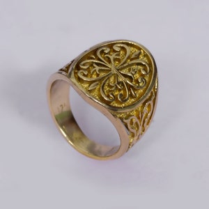 May include: A gold-colored ring with an oval face featuring an intricate filigree design. The ring's band is smooth, and the face has a raised, detailed pattern. The ring is likely made of gold or a gold-toned metal, showcasing a vintage or antique style.