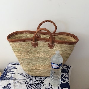 May include: A large, brown leather-trimmed straw tote bag with two handles. The bag is sitting on a white tablecloth with a blue and white pattern. A clear plastic water bottle is sitting next to the bag.
