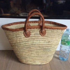 May include: A large, natural woven basket with brown leather handles and trim. The basket is sitting on a wooden floor next to a clear plastic water bottle.