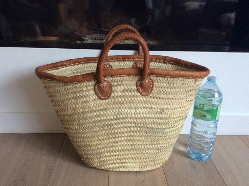 May include: A large, natural woven basket with brown leather handles and trim. The basket is sitting on a wooden floor next to a clear plastic water bottle.