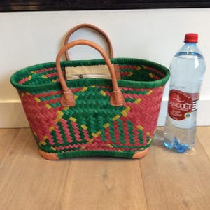 May include: A green, pink, and yellow woven basket bag with brown leather handles. The bag is sitting on a wooden floor next to a clear glass bottle of water.