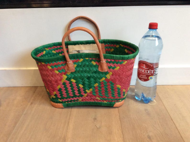 May include: A green, pink, and yellow woven basket bag with brown leather handles. The bag is sitting on a wooden floor next to a clear glass bottle of water.