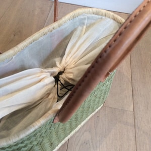 May include: A green woven basket with brown leather handles and a white cotton lining. The lining is gathered at the top and secured with a black drawstring.