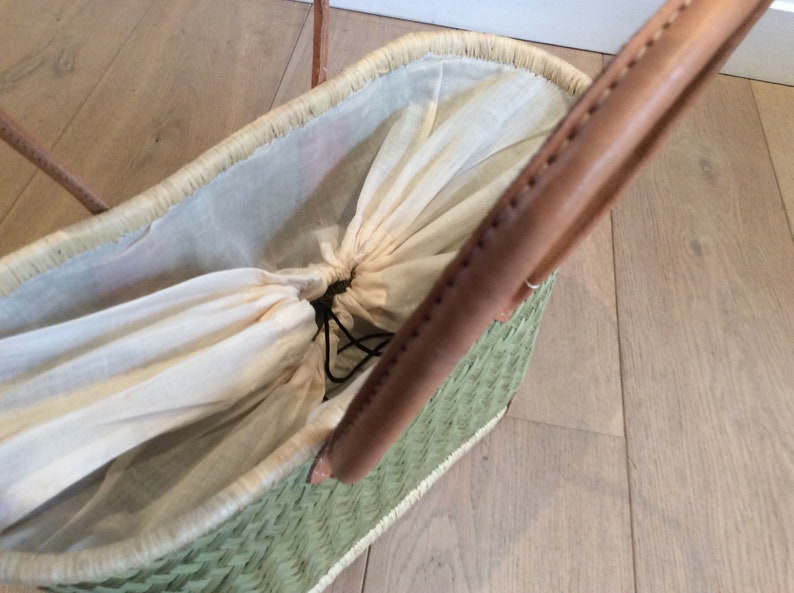 May include: A green woven basket with brown leather handles and a white cotton lining. The lining is gathered at the top and secured with a black drawstring.