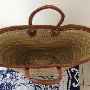 May include: A large, oval-shaped straw tote bag with brown leather handles and trim. The bag is empty and the inside is visible.