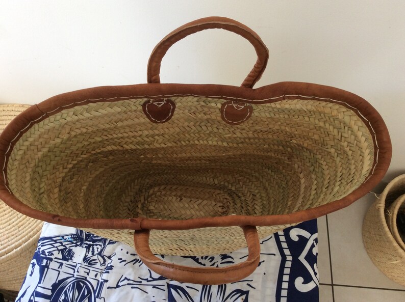 May include: A large, oval-shaped straw tote bag with brown leather handles and trim. The bag is empty and the inside is visible.