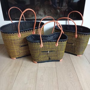 May include: Three woven tote bags in different sizes. The bags feature a yellow and brown woven pattern, black lining, and tan leather handles. They are displayed on a light-coloured wooden floor.
