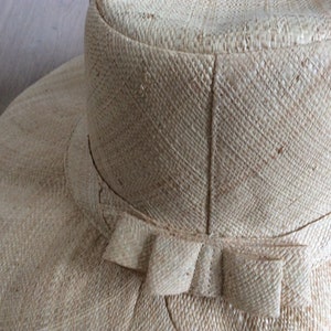 Puede incluir: Un sombrero de paja de ala ancha con un gran lazo en la parte posterior. El sombrero est&aacute; hecho de un material natural tejido y tiene un color beige claro.
