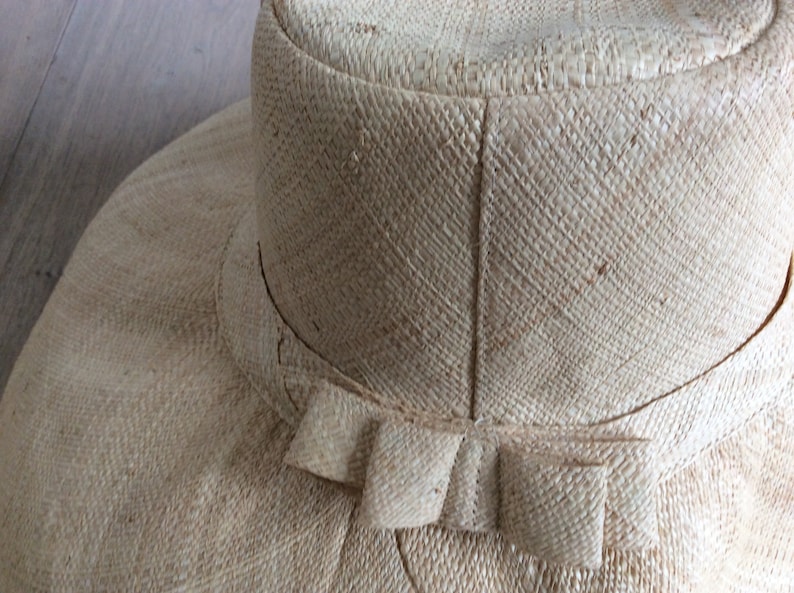 Puede incluir: Un sombrero de paja de ala ancha con un gran lazo en la parte posterior. El sombrero est&aacute; hecho de un material natural tejido y tiene un color beige claro.