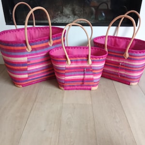May include: Three pink and striped tote bags of different sizes. The bags have light brown handles and are made of woven material. The stripes include shades of pink, orange, purple, and white. The bags are on a light wood floor.