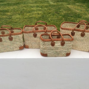May include: Set of five woven straw tote bags with brown leather handles and trim. The bags are arranged in a row from largest to smallest.