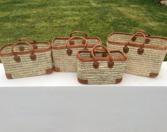Puede incluir: Conjunto de cinco bolsas de mano de paja tejidas con asas y ribetes de cuero marrón. Las bolsas están dispuestas en fila de la más grande a la más pequeña.