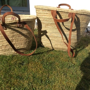 May include: Two woven straw baskets with leather handles. The larger basket is on the right and the smaller basket is on the left. Both baskets are sitting on a green grassy surface.
