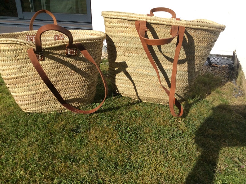 May include: Two woven straw baskets with leather handles. The larger basket is on the right and the smaller basket is on the left. Both baskets are sitting on a green grassy surface.