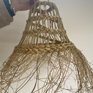 May include: A woven lampshade made of natural materials, with a bell shape and a fringe of long strands. The lampshade is light brown in color and has a rustic, handmade look.