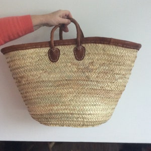 May include: A large, natural woven straw tote bag with brown leather handles and trim. The bag has a wide, open top and a rounded bottom.