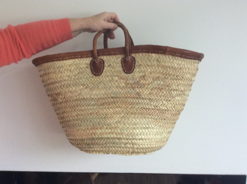 May include: A large, natural woven straw tote bag with brown leather handles and trim. The bag has a wide, open top and a rounded bottom.