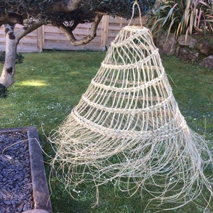 Op de afbeelding: Een grote, geweven, lichtbruine lampenkap hangt aan een tak in een tuin. De lampenkap is gemaakt van natuurlijke vezels en heeft een gefranst uiteinde.