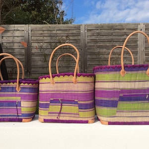 May include: Three woven tote bags with leather handles. The bags are striped in shades of purple, green, and beige. The largest bag is in the back, followed by a medium-sized bag, and a small bag in the front.