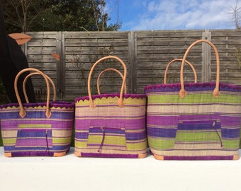 May include: Three woven tote bags with leather handles. The bags are striped in shades of purple, green, and beige. The largest bag is in the back, followed by a medium-sized bag, and a small bag in the front.
