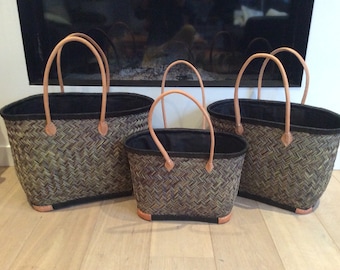 May include: Three woven baskets with black fabric lining and brown leather handles. The baskets are different sizes and are arranged in a row on a wooden floor.