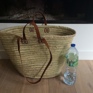 May include: A large, oval-shaped, woven straw basket with brown leather handles. The basket is sitting on a wooden floor next to a clear plastic water bottle.