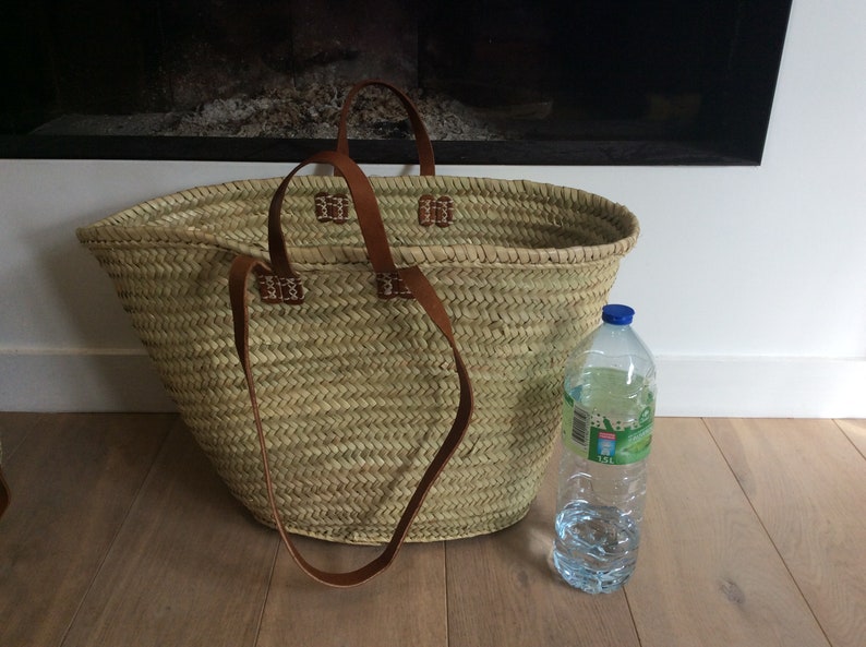 May include: A large, oval-shaped, woven straw basket with brown leather handles. The basket is sitting on a wooden floor next to a clear plastic water bottle.
