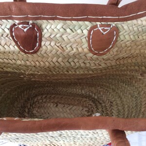 May include: A woven straw tote bag with brown leather handles and trim. The bag has two brown leather patches on the inside with white stitching.
