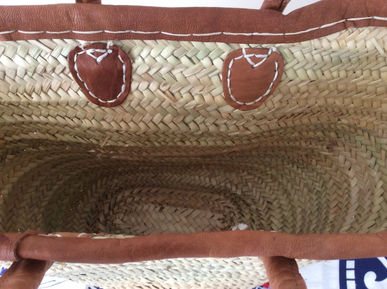 May include: A woven straw tote bag with brown leather handles and trim. The bag has two brown leather patches on the inside with white stitching.