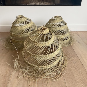 Op de afbeelding: Drie natuurlijke geweven lampenkappen gemaakt van stro met een lichtbruine kleur. De lampenkappen zijn rond en hebben een uniek, ingewikkeld ontwerp.