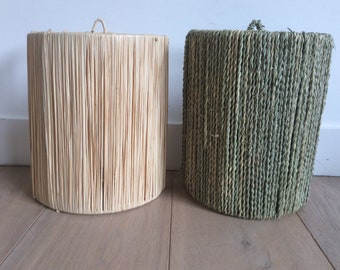 May include: Two cylindrical woven lampshades, one in natural beige and the other in green. Both shades are made of a natural fiber material, possibly seagrass or rattan.