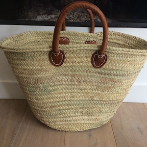 May include: A large, oval-shaped woven basket with brown leather handles. The basket is made of natural straw and has a light brown colour.