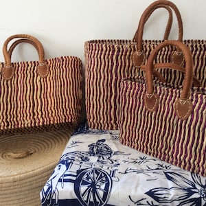 Puede incluir: Tres bolsas de mano de paja tejidas con asas de cuero. Las bolsas están rayadas en tonos beige y morado. La bolsa más grande está en la parte posterior, y la bolsa más pequeña está en la parte delantera.