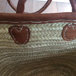May include: A woven straw tote bag with brown leather handles and trim. The bag has two brown leather patches on the inside with white stitching.