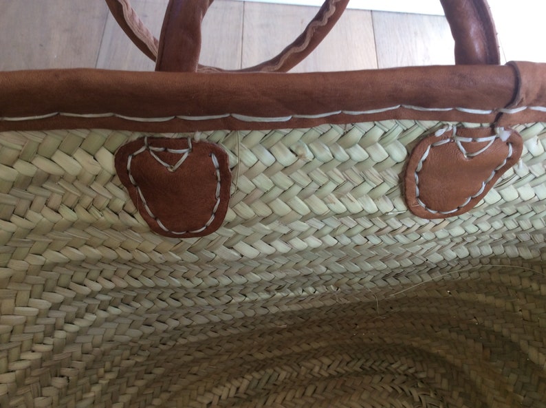 May include: A woven straw tote bag with brown leather handles and trim. The bag has two brown leather patches on the inside with white stitching.