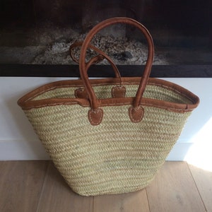 May include: A large, woven straw tote bag with brown leather handles and trim. The bag is light brown and has a slightly rounded shape.
