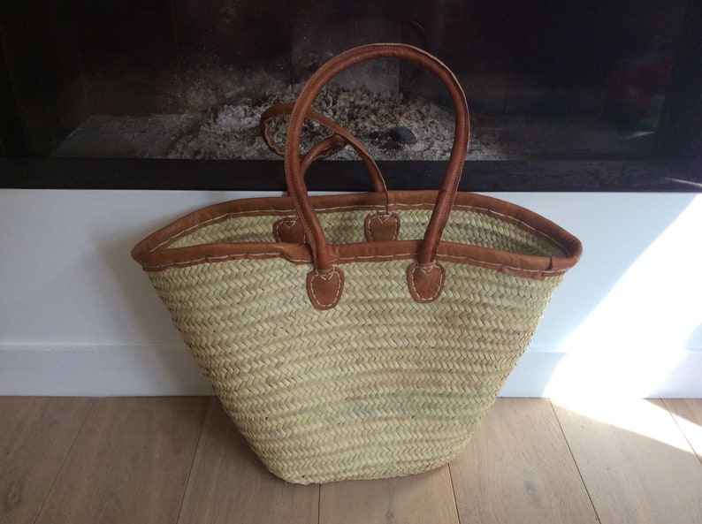 May include: A large, woven straw tote bag with brown leather handles and trim. The bag is light brown and has a slightly rounded shape.