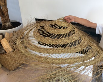 Puede incluir: Una pantalla de lámpara grande, redonda y tejida hecha de materiales naturales. La pantalla de la lámpara es marrón y tiene un aspecto natural y rústico. Está siendo sostenida por la mano de una persona.