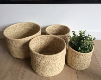 Puede incluir: Conjunto de cuatro cestas tejidas en color beige natural. Las cestas son redondas y tienen una textura natural tejida a mano. La cesta más grande está en primer plano, con las otras tres cestas apiladas detrás.