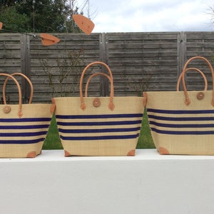 Könnte beinhalten: Drei gestreifte Einkaufstaschen aus Naturfaser mit Ledergriffen und Besätzen. Die Taschen sind der Größe nach aufgereiht, die größte Tasche links und die kleinste Tasche rechts. Die Taschen sind alle mit blauen und naturfarbenen Streifen versehen.