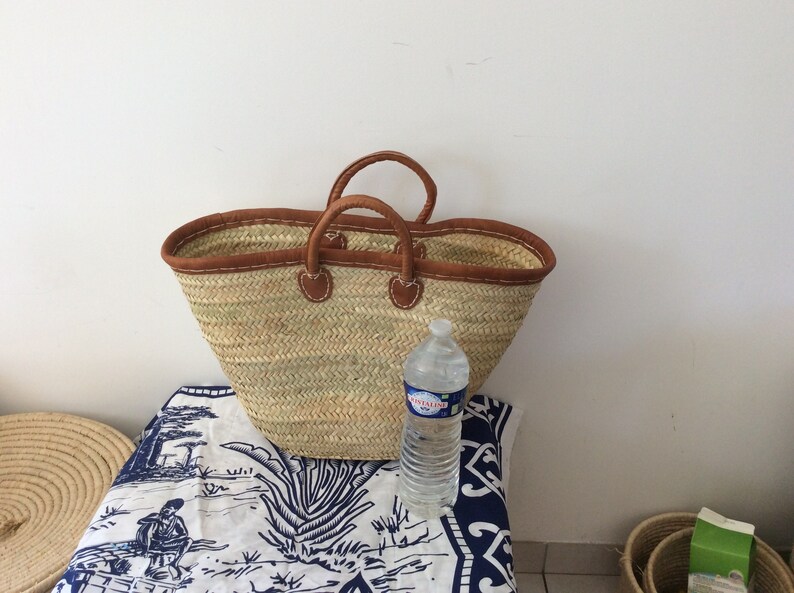 May include: A large, brown leather-trimmed straw tote bag with two handles. The bag is sitting on a white tablecloth with a blue and white pattern. A clear plastic water bottle is sitting next to the bag.