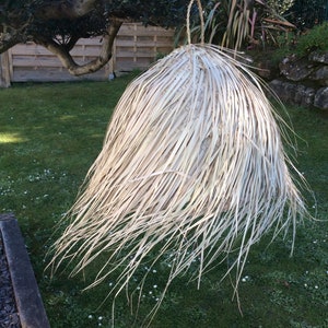 Op de afbeelding: Een grote, witte, geweven lampenkap gemaakt van natuurlijke materialen hangt in een grasrijke buitenomgeving.