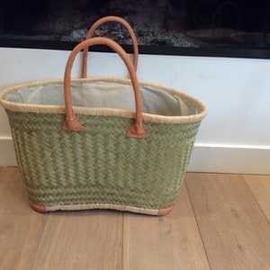 May include: A green woven basket with leather handles. The basket is oval shaped and has a white lining.
