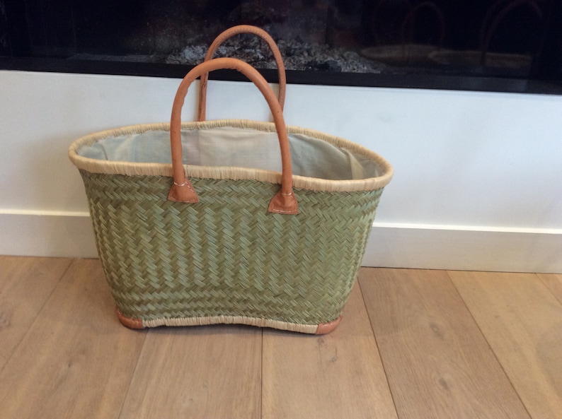 May include: A green woven basket with leather handles. The basket is oval shaped and has a white lining.