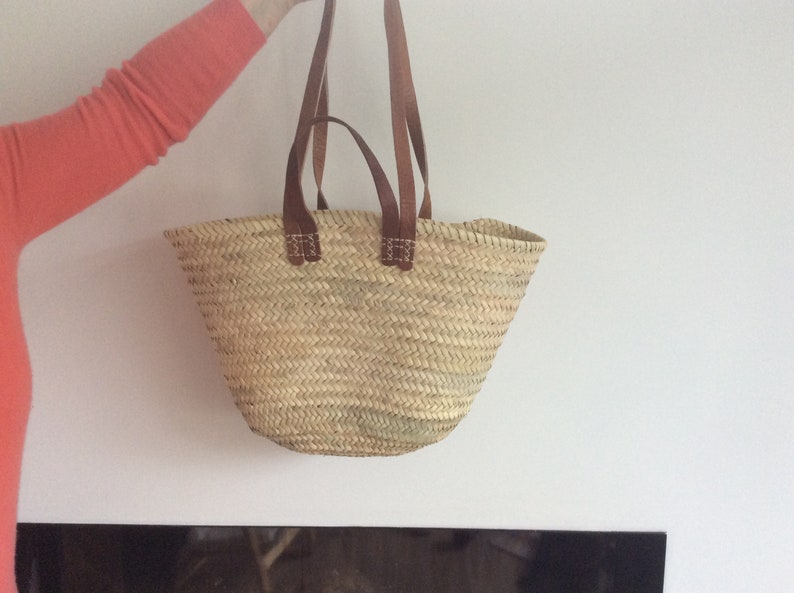 Puede incluir: Un gran bolso de mano de paja trenzada natural con asas de cuero marr&oacute;n. El bolso est&aacute; colgado de las asas y tiene una parte superior ancha y abierta.