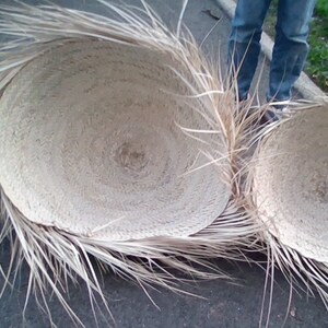 Op de afbeelding: Twee grote, ronde, geweven manden met een natuurlijke, lichtbruine kleur. De manden zijn gemaakt van een natuurlijk materiaal, mogelijk palmbladeren, en hebben een gefranst randje. De manden staan op een grijs oppervlak.